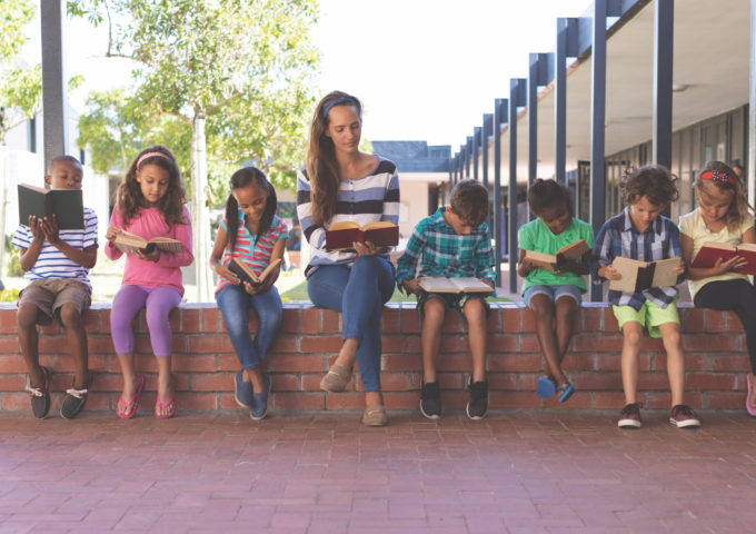 Front view of caucasian teacher and multi ethnic students reading book while sitting on brick wall at corridor in school