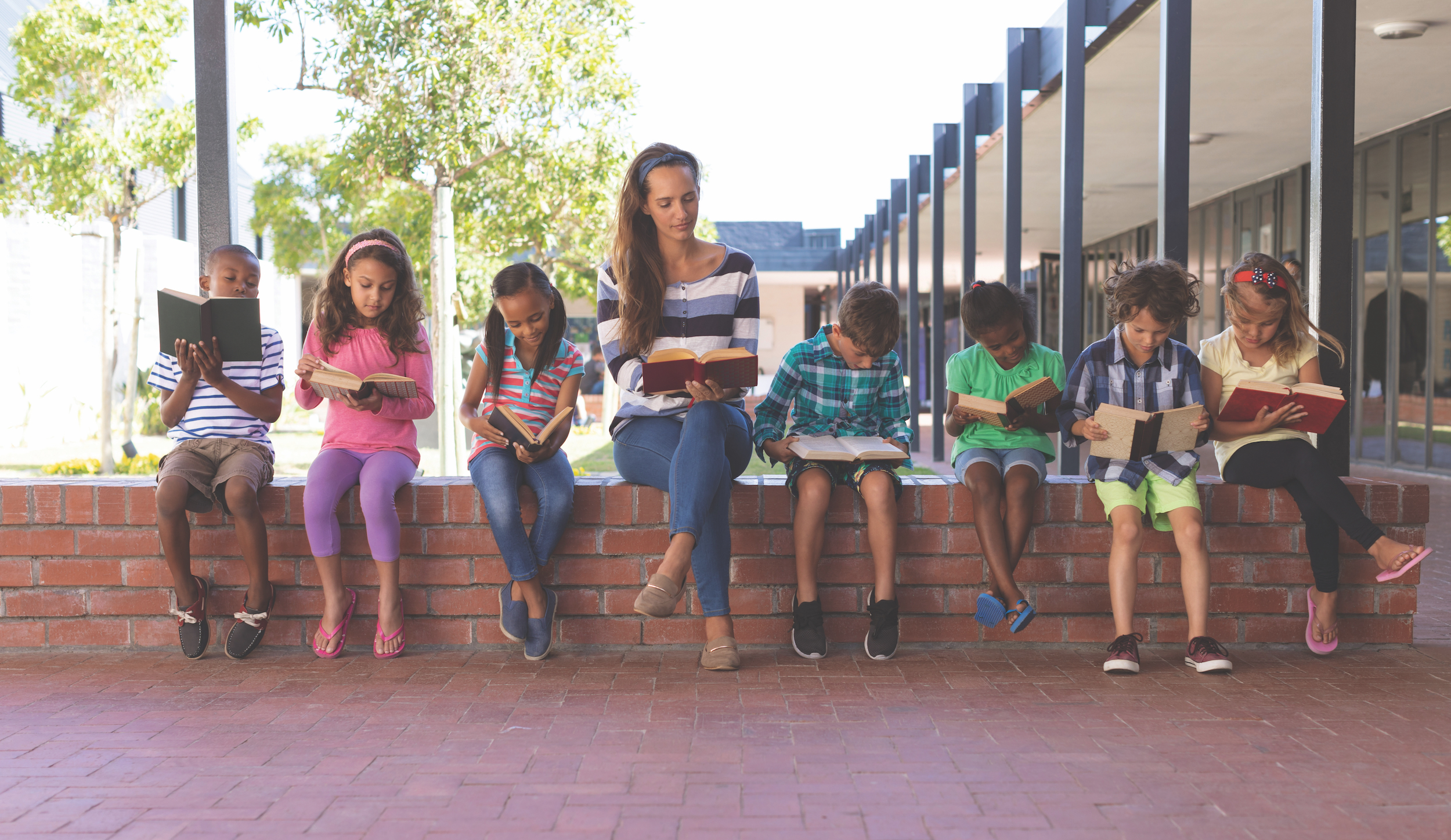 Front view of caucasian teacher and multi ethnic students reading book while sitting on brick wall at corridor in school