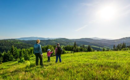 Rear view young mother and two daughters go down the hill overgrown with green grass. Gorgeous view of the forest growing on the mountains during trekking on a warm summer day