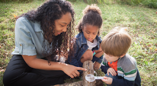 Woman uses a magnifying glass with two young children to inspect details on a log.