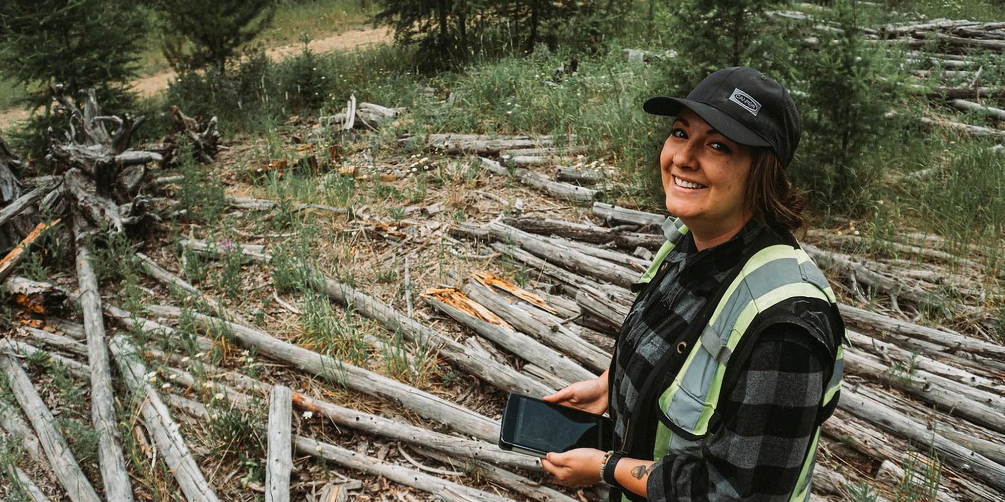 Woman with a tablet computer and high vis vest standing in front of some old tree trunks on the ground