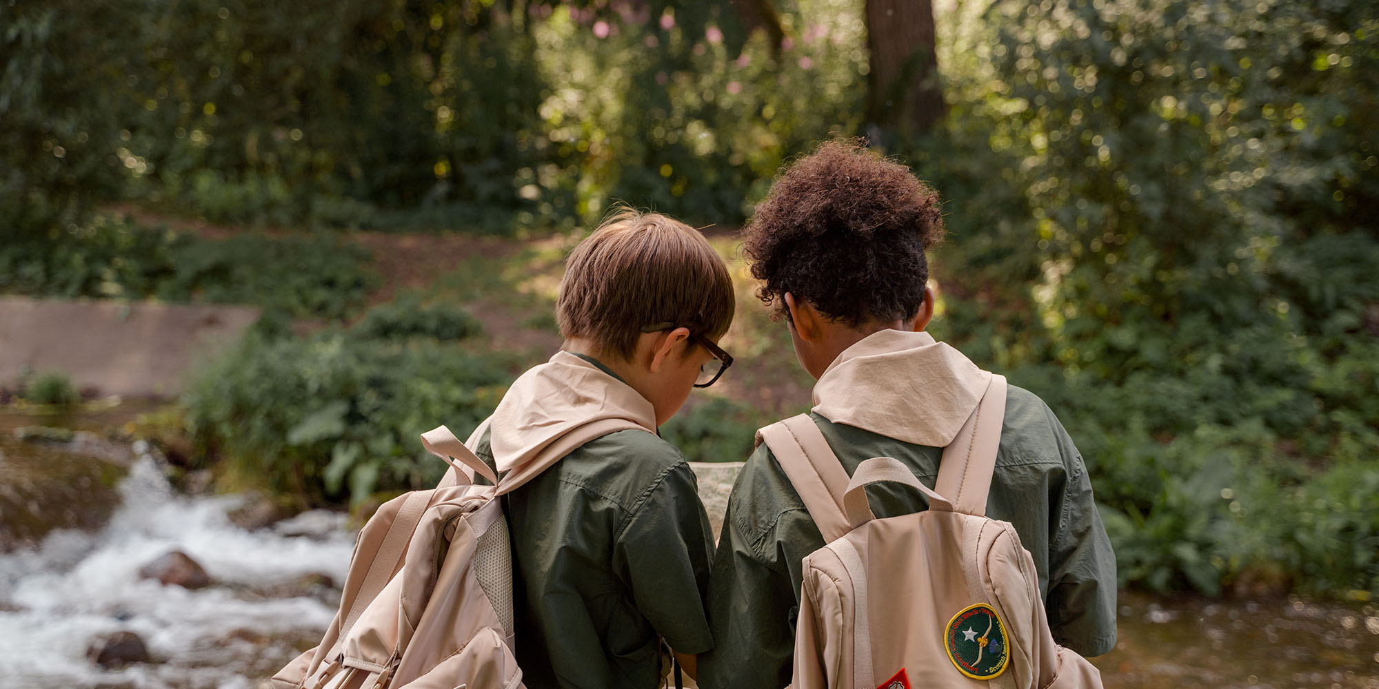 Two young boys in scouts uniforms look at a map together in the forest.