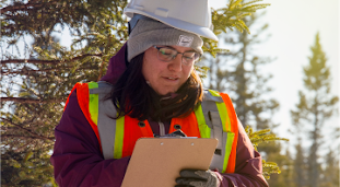 A woman in a hard hat and high visibility vests stands in a forest and takes notes on a clip board.