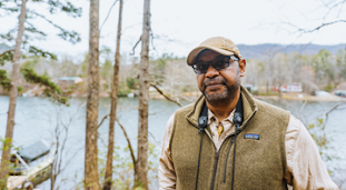 A man in a vest and ball cap stands in a forest in front of river or lake